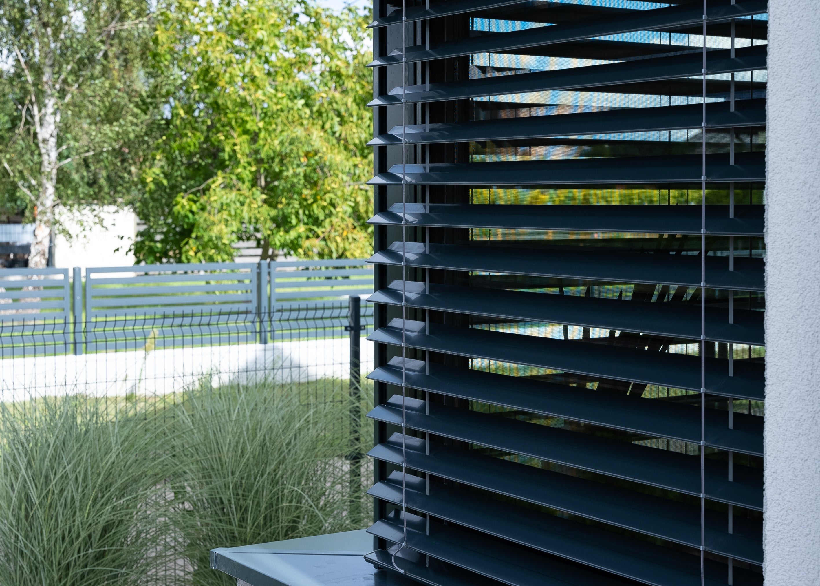 Modern architectural detail of a home with exterior venetian blinds and greenery in the background